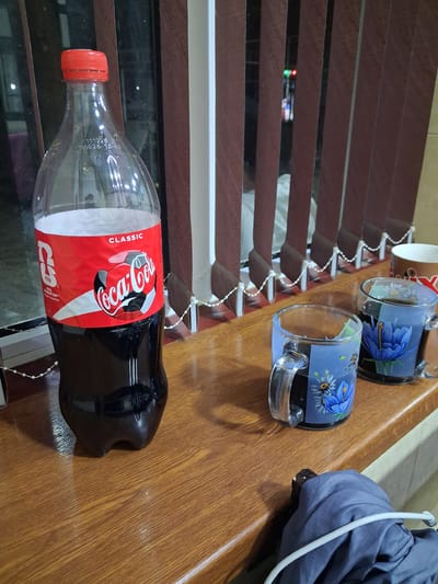 The image shows a Coca-Cola bottle and two decorative glass mugs on a wooden surface. The Coca-Cola bottle is red and features the iconic Coca-Cola logo along with "CLASSIC" and FIFA World Cup branding. The two glass mugs are decorated with blue flowers and green leaves, and appear to contain a dark liquid. A bundle of gray fabric, possibly clothing, is also visible on the right side of the frame, with a white cord draped over it.
In the background, horizontal wooden blinds are visible, and behind them, a glimpse of a surface with red and green lights can be seen, suggesting some kind of electronic device. The lighting in the image is warm, indicating it was likely taken indoors during the day. The composition of the image suggests a casual setting, perhaps a home or a casual workspace. The Coca-Cola bottle seems to be the main focus, with the other items serving as supporting elements in the scene. The visible text confirms the product and a promotional event related to the FIFA World Cup.