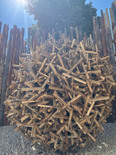 The image shows a large, spherical sculpture made of numerous pieces of light-colored wood, arranged in a seemingly random yet structured manner. The sculpture is situated outdoors, resting on a bed of light gray gravel. Behind the sculpture, a rustic fence constructed from vertical wooden poles of varying thickness and bark texture encloses the area. The sky above is a clear blue with bright sunlight streaming down, creating lens flare and casting shadows. A dense green tree with dark foliage is visible behind the wooden fence, partially obscuring the sky. The overall setting appears to be an outdoor exhibition or art installation. The image does not contain any people or identifiable animals.