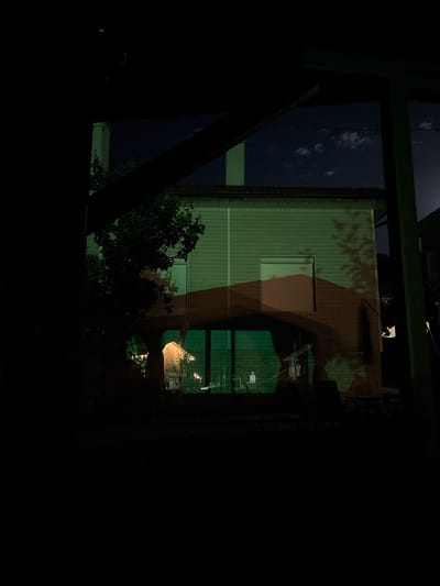 The image shows a view from inside a dark space, framed by a dark green structure, likely part of a building or industrial framework. Through this opening, a building with vertical siding is visible, illuminated by artificial light. The building's facade appears to have a window and is partially covered by shadows, possibly cast by tree branches. To the left, a dark blue area suggests a night sky. The lower part of the view is obstructed by dark foliage. The city of Polena, Bulgaria is not discernible in the image.