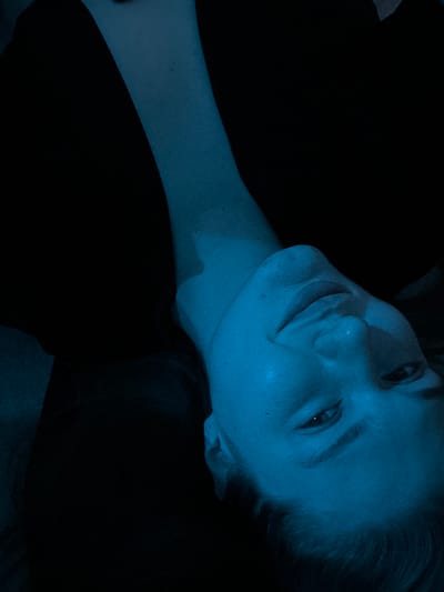 A person with fair skin is lying down. The head is tilted to the left, and the person is smiling slightly. The neck extends towards the upper right corner. The background is mostly dark, with some dark material behind the head and around the neck. The overall image has a blue cast. The lighting is dim, creating strong shadows.