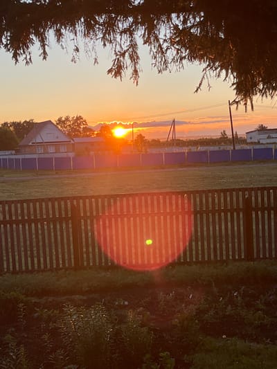 The image depicts a rural landscape at sunrise, with the sun casting a warm orange glow. In the foreground, a wooden fence runs horizontally across the frame, separating the viewer from a grassy area and a garden with lush green plants. Beyond the fence, a blue fence encloses a property with a two-story house. The house is light-colored with a brown roof and white-framed windows. In the background, trees and power lines are visible against a sky filled with soft clouds and the rising sun. The overall scene is peaceful and suggests a typical morning in a Russian village. The location is identified as Baimovo, Russia.