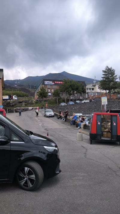 A cloudy, overcast day is depicted at the Rifugio Sapienza on Mount Etna, near Nicolosi, Italy, a popular starting point for volcano excursions. In the foreground, the front left portion of a black Mercedes-Benz minivan is visible, parked on an asphalt surface. To the right of the minivan, a red and black cable car cabin is parked, appearing to be out of service. Behind this, a group of people are seated on a low stone wall, some looking towards the volcano or interacting with each other. Further back, several cars are parked in a lot. A building with a large sign that reads "FUNIVIA DELL'ETNA" (Etna Cableway) is prominently featured, with ski lifts ascending the barren, volcanic slope of Mount Etna in the background. The environment is characterized by volcanic rock, sparse vegetation, and a general feeling of a high-altitude, potentially weathered location.