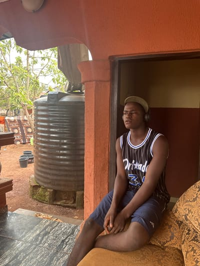 The scene depicts a young Black male seated on a patterned sofa in a semi-enclosed porch or doorway within Agbogugu, Nigeria. The male wears a light green cap, black over-ear headphones, a black and white striped basketball jersey with "Orlando 32" printed on it, and dark blue shorts. His hands are clasped, and he gazes forward with a neutral expression.
To his left, the immediate outdoor environment features a large, dark grey ribbed plastic water storage tank resting on a stone base. Beyond the tank, several black plastic buckets are visible on reddish-brown earth. A patterned fabric item hangs in the background to the left of the tank. The surrounding area exhibits lush green foliage under an overcast sky. The architectural structure consists of orange-red painted pillars and an archway, framing the view to the exterior. The floor beneath the male's feet appears to be dark, possibly black, marble-patterned tile.
