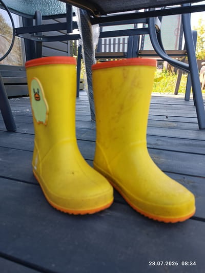 A pair of yellow children's rubber boots with orange soles and tops are displayed on a dark wooden deck. The left boot features a light green cartoon duck emblem with an orange outline. The boots are positioned slightly angled, with the left boot closer to the viewer. In the background, the legs of outdoor furniture, a grassy area, and trees are visible. The scene is set outdoors, likely in a residential yard, with no specific indication of the city of Stavropolskaya, Russia. The lighting suggests it is daytime. A timestamp "28.07.2026 08:34" is present in the bottom right corner of the image.