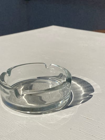 A clear, circular glass ashtray is centrally positioned on a white, textured surface, consistent with painted wood. The ashtray features four distinct cigarette rests along its rim and exhibits minor dark particulate residue in its base. Strong, direct sunlight illuminates the scene, casting a prominent, ribbed shadow from the ashtray onto the surface to its right. A broad, diagonal shadow from an unseen overhead structure or object bisects the upper background, extending across the top-left portion of the white surface. No individuals, actions, or interactions are present. The location is Πλατύς Γιαλός, Greece.