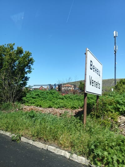 The image shows a road sign indicating the entrance to Venets, Bulgaria. The sign is white with black Cyrillic and Latin script. Behind the sign, there is lush green vegetation and trees. In the background, there are buildings with metal roofing and a cell tower on a hill. The sky is clear blue. The photograph appears to have been taken from a moving vehicle.