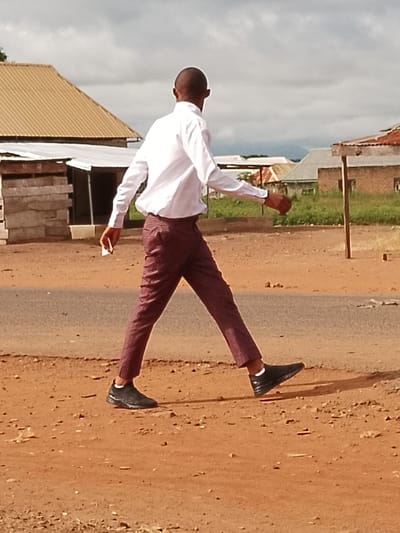 A young man walks along a dirt road in Ichol, Nigeria, wearing a white long-sleeved shirt and patterned maroon pants. He is holding a white object in his right hand. The background shows several buildings with corrugated metal roofs, some appearing unfinished, and a cloudy sky. Green vegetation is visible behind the structures. The road is unpaved, with the surface appearing dry and dusty.