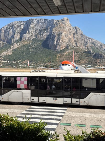 The image shows an airport scene, possibly in Cinisi, Italy, given the mountainous backdrop typical of the region. An airplane with orange and white livery is parked near a passenger bus, with a mountain visible in the background. The bus is on a striped crosswalk on an airport apron. The foreground includes some foliage.