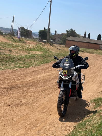 A motorcyclist wearing a helmet and dark clothing is riding a white, red, and black motorcycle on a dirt road. The motorcycle has a number "5" sticker on the front. Behind the rider is a building with a tiled roof and tall, slender trees. In the background, there is a view of a cityscape and rolling hills. A banner with the word "AHAMAY" is visible on a pole to the left. The photo was taken in the city of l'Ametlla del Vallès, Spain.