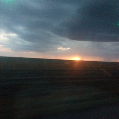 A wide, horizontal, slightly out-of-focus image captures a sunset over a vast, flat, dark expanse, likely a field or plain, near Adyk, Russia. The sky is overcast with dark, heavy clouds dominating the upper portion, transitioning to lighter, muted tones near the horizon. A bright, intense spot of sunlight is visible on the horizon, partially obscured by a thin band of clouds and a small, fluffy cloud directly above it, indicating the sun is either setting or has just risen. The foreground is a dark, indistinct mass, suggesting a large, flat landscape with very little visible detail. No people, structures, or distinct objects are present in the scene. The overall impression is one of a desolate, natural landscape at twilight.