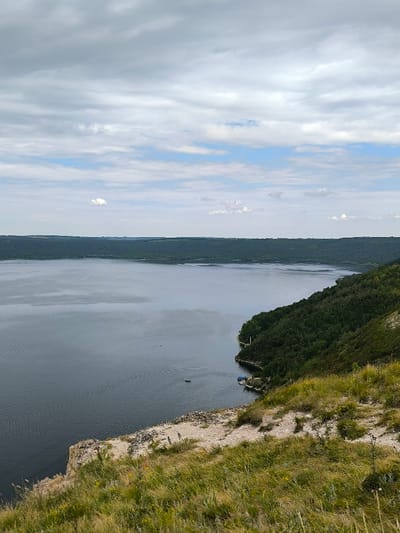 The image displays an elevated, panoramic view of a wide water body, identified as the Dniester River reservoir, observed from a cliffside near Horaivka, Khmelnytskyi Oblast, Ukraine. The upper portion of the frame is occupied by a sky showing a mix of stratified grey and white clouds with visible patches of clear blue.
The dark grey water of the reservoir fills the central and lower-left sections of the image. In the immediate foreground, a downward-sloping terrain comprises dry, light-colored soil and sparse, mixed green and straw-colored grass, leading to a prominent rocky cliff edge. The right bank of the reservoir descends sharply from this foreground, covered densely in dark green deciduous forest that extends to the water's edge. A small, light blue structure, consistent with a boathouse or shelter, is situated at the base of this heavily vegetated bank. A small, dark-colored boat is moored adjacent to this structure.
Approximately one-third from the left edge and one-third from the bottom, a small, light-colored boat is visible on the water, generating a distinct V-shaped wake indicating forward movement. The opposing bank across the reservoir is also densely forested with dark green foliage, forming a relatively uniform, elevated ridge against the distant horizon. No human figures are identifiable within the scene.