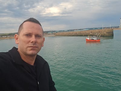 A man in a black jacket and t-shirt poses for a selfie on a pier overlooking a body of water. The water is a murky green color and appears to have moderate chop. In the distance, a small orange and white fishing boat is visible. A long concrete pier stretches across the water, with buildings and a beach visible beyond it. The sky is overcast with dramatic clouds. The scene is likely located in Shoreham-by-Sea, within the Adur district of West Sussex, United Kingdom, given the presence of the fishing boat and harbor structures commonly found in the area.