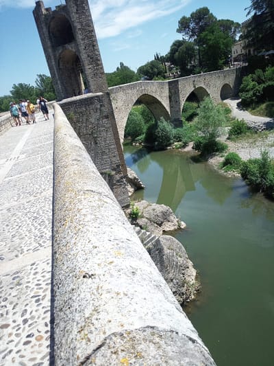 The image shows the medieval bridge of Besalú, Spain, spanning the Fluvià River. The bridge is constructed of stone and features several arches. A large, fortified tower with crenellations stands at the entrance to the bridge, leaning at an angle. Several people are walking across the bridge's cobblestone path, appearing to be tourists. The river below is a murky green, reflecting the stone structure of the bridge. Lush green trees and vegetation line the riverbanks and the surrounding landscape. The sky is blue with scattered white clouds. The perspective of the image is from a low angle, looking up at the bridge and tower, with the camera positioned on the bridge's stone railing.