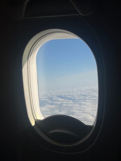 The image is a view from an airplane window, looking out at a sky filled with white clouds and a bright blue sky above. The window frame is dark, with a white, curved interior. The image does not contain any information about the city of Kczewo, Poland.