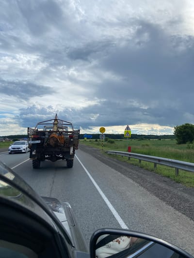 This media file depicts a view from a car driving on a road in Bagrash-Bigra, Russia. The primary subjects are the road, a vehicle in front, a guardrail, and the surrounding landscape under a dramatic sky.

The vehicle in front appears to be a specialized agricultural or industrial machine, possibly a sprayer or a similar piece of equipment, with a prominent boom structure. It has large tires and a weathered appearance, with visible license plates. Following closely behind is a white passenger car. The perspective from inside the car shows a side mirror reflecting a person wearing a light-colored shirt with a graphic design.

The setting is a rural or semi-rural area. The road is paved with a white lane marker. To the right of the road, there is a metal guardrail and a grassy verge, beyond which stretches a green field with some trees in the distance. To the left, the sky is filled with heavy, dark clouds, suggesting impending rain or a recent storm, but there are also patches of brighter sky and sunlight breaking through, creating a dynamic and atmospheric scene. Several road signs are visible, including a triangular yellow sign with a symbol and a circular yellow sign.

The activity taking place is travel by road. The time of day appears to be daytime, given the natural light. The weather is cloudy and possibly moody, contributing to the dramatic visual.

A license plate on the rear vehicle is partially visible, reading "A 738 AO 18". Another road sign in the distance displays some blue text, which is not legible. The overall impression is of a journey through the Russian countryside under a striking sky.