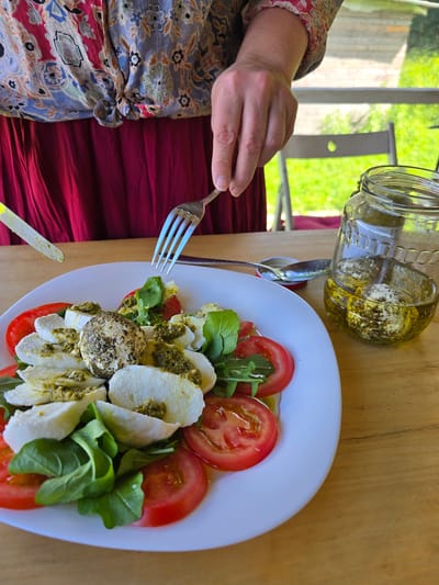 A person's hands are visible in the frame, one holding a fork and about to interact with a caprese salad. The salad is arranged on a white plate and features slices of fresh mozzarella, ripe tomatoes, and green arugula, all drizzled with a vibrant green pesto. A glass jar filled with cheese and oil is positioned in the upper left corner, hinting at the preservation of ingredients. The scene appears to be set outdoors, possibly on a patio or in a garden, with lush greenery visible in the background, suggesting daytime and pleasant weather. The setting suggests a meal being enjoyed in a relaxed, possibly rural, environment, with the presence of fresh ingredients and a casual dining setup. The wooden table surface provides a rustic touch to the scene. No visible text is present in the image.
