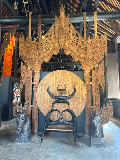 A complex interior features elaborate wooden carvings, reminiscent of wings, attached to black columns. A large, circular wooden panel carved with intricate designs is prominently displayed, overlaid with stylized, curved black horns. Above the panel, there is a sculpture of a horse's head. Below it, there is another horse's head sculpture and a framed print. The setting evokes a museum or gallery space.