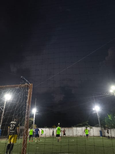 This image depicts a nighttime futsal or soccer match on an artificial turf field in Timaná, Colombia. The foreground is dominated by the upright of a goal post and its net, behind which a player wearing a black and yellow jersey with "MURCIA 27" on the back stands. A protective black netting extends across the entire upper portion of the frame, obscuring the dark, cloudy sky.

On the illuminated green artificial turf field, approximately ten male individuals are present, participating in the game. Players wear various colored jerseys, including prominent lime green, dark blue, black, and red. They are spread across the field, with some in various stances of play. The field is bordered by a low, white concrete wall or fence. Bright floodlights on poles illuminate the scene from the right and left sides, casting strong light onto the field. Beyond the perimeter wall, dark vegetation, possibly trees or bushes, is visible under the intense artificial light. One individual is discernible standing just beyond the far wall. The overall environment is an outdoor sports facility active at night.
