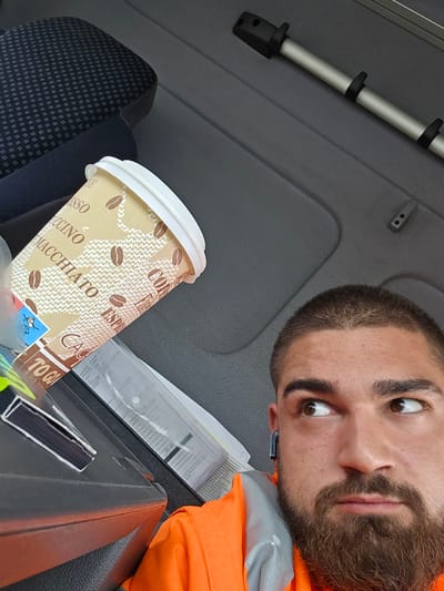 A close-up, low-angle shot depicts a man with a beard and an orange vest, seemingly lying down inside a vehicle. A coffee cup with coffee bean designs sits on the vehicle's dashboard. A gray-colored dashboard surface, parts of a car seat, and a metallic pole are visible as well. The exact location in Halle (Saale), Germany, cannot be determined with the information provided. No specific actions or interactions are discernible.