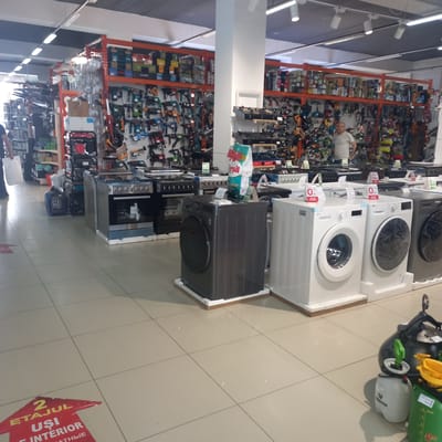 This is an image of the interior of a store, likely a home improvement or appliance store, located in Briceni, Moldova. The store is well-lit with overhead fluorescent lighting. The floor is tiled in a light beige color.

In the foreground, on the right side of the image, are several cleaning appliances. There is a large black appliance, possibly a steam cleaner, with a green spray nozzle attached. Next to it is a white spray bottle labeled "VERTO" and a green jug labeled with measurements and "max". Further to the left, in the center and extending to the left, are several washing machines. There is a dark grey washing machine, followed by two white washing machines. All appear to be new and on display, some still with plastic wrap on the doors. Behind the washing machines, in the middle ground, are various kitchen appliances, including gas stoves and ovens, arranged in rows.

The background of the image shows large shelving units filled with a variety of merchandise, predominantly tools and hardware. These shelves are orange metal structures. On the shelves, one can see numerous power tools, such as drills and sanders, as well as boxes of various sizes and colors, likely containing hardware, electrical supplies, or other DIY items. Some of the boxes are labeled with brand names.

A man wearing a light-colored t-shirt is visible in the mid-ground, standing near the shelving units, looking towards the right. To the far left, a person is walking away from the camera, carrying a white plastic bag.

On the floor in the lower left corner of the image, there is a red directional arrow sticker with text in what appears to be Romanian, indicating "2 ETAJUL UȘI INTERIOR", suggesting an upstairs department for interior doors.

The overall impression is of a busy retail environment offering a wide range of household and construction goods.