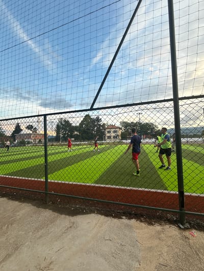 The image displays an artificial turf football or soccer field viewed through a protective mesh fence. The fence, composed of a dark metal frame and fine netting, runs across the foreground, slightly obscuring the view of the field.
On the bright green artificial pitch, marked with white lines and a reddish-brown border, several individuals are present, suggesting a game or practice session. In the midground, two men are more clearly visible: one wears a neon yellow shirt and black shorts, while the other is dressed in a blue shirt and red shorts. Both appear to be engaged in activity or moving across the field. Other figures can be seen further down the pitch.
Beyond the field, a background of lush green trees and a white multi-story building with dark windows indicates a developed area. Distant hills or mountains are faintly visible under a partly cloudy sky. The lighting suggests it is late afternoon or early evening. To the right of the fence, a concrete and dirt path borders the playing area. No legible text is visible within the frame. The scene is located in Blatska, Bulgaria.