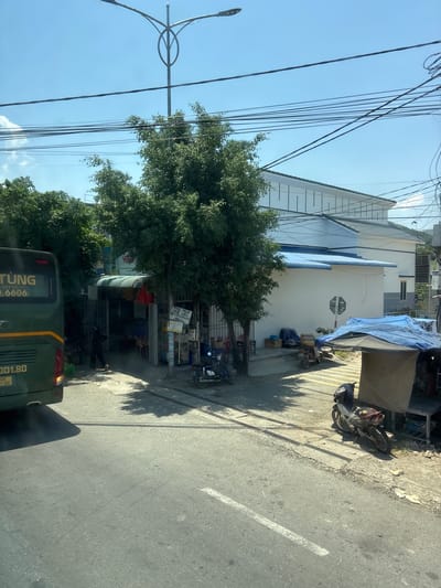 A wide shot captures a sunlit street scene with a dark green bus on the left, partially obscuring a small roadside shop. The bus has text on its side including "TUNG" and "001.80". The shop appears to have a corrugated metal roof and a narrow entrance. Trees with dense green foliage are prominent in the foreground, casting shadows. Beyond the shop, a modern white building with a blue roof is visible, characterized by a clean facade and numerous horizontal white bars. Power lines crisscross the bright blue sky, a common sight in urban environments. In front of the white building, a covered stall with a tarp is set up, sheltering some motorcycles and miscellaneous items. A few parked motorcycles are visible on the right side of the image, some under tarps. The road in the foreground is asphalt with a white line marking a lane. The overall impression is one of a bustling, everyday street in a populated area, likely in Southeast Asia, given the architecture and the presence of motorbikes.