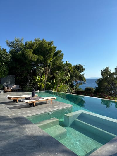 This image depicts an outdoor infinity pool with a view of the sea, likely located in the Vouliagmeni Municipal Unit, Greece. Two wooden lounge chairs with cushions and towels are positioned on a grey tiled patio. A black speaker sits on one of the lounge chairs. The pool itself is filled with clear turquoise water and features built-in steps. Lush green trees and foliage surround the pool area, and in the distance, a yacht is visible on the calm blue sea under a clear sky. The overall scene suggests a luxurious and serene vacation setting.