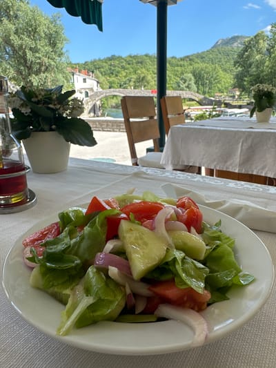 The image shows a close-up of a fresh salad on a white plate, placed on a white tablecloth. The salad consists of lettuce, sliced tomatoes, cucumber, and red onion. In the background, a picturesque scene unfolds in Rijeka Crnojevića, Montenegro. A stone bridge with multiple arches spans a calm body of water, with traditional stone buildings visible on the far bank. Lush green hills covered in trees form the backdrop under a clear blue sky. A dark metal pole with a green awning is partially visible in the foreground, suggesting an outdoor seating area of a restaurant. Tables with white tablecloths and chairs are also present, indicating a dining setting. A small potted plant with white flowers sits on a table to the right, and a glass bottle containing red liquid, likely oil or vinegar, is on the left. The overall impression is one of a tranquil dining experience in a scenic historical location.