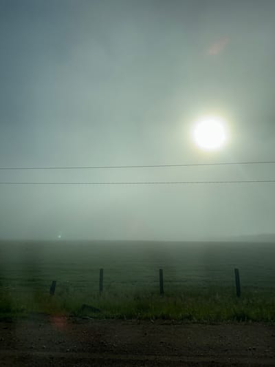 The image depicts a foggy rural landscape, possibly in or near Kuda, Russia, based on the provided context. In the foreground, there is a dirt road with sparse green grass along its edge. Several wooden fence posts are visible protruding from the ground, spaced irregularly across the field. The field itself is covered in green grass and stretches into the distance, where it is obscured by a thick layer of fog. In the sky, a bright, hazy sun is positioned in the upper right quadrant, its light diffusing through the atmospheric conditions. Two parallel power lines are stretched horizontally across the frame, separating the sky from the land. The overall atmosphere is muted due to the fog, with the sun creating a bright, diffused glow. No people or animals are present in the scene.