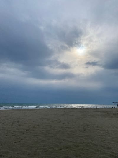 The image presents a wide view of a sandy beach in Ulcinj, Montenegro, under an overcast sky. The foreground comprises a textured expanse of light brown sand. The midground reveals the ocean with visible waves breaking along the shore, exhibiting a dark teal-blue coloration. A strong, silvery reflection of a diffuse light source, likely the sun obscured by clouds, extends across the water's surface towards the horizon. The upper two-thirds of the frame are dominated by dense, dark grey cloud cover, with a concentrated area of brighter light visible in the upper center-right. On the far right edge of the beach, a partial wooden structure, consisting of vertical posts and a horizontal crossbeam, is visible. Several slender, dark vertical poles protrude from the water farther offshore. No individuals or other significant objects are present in the scene.