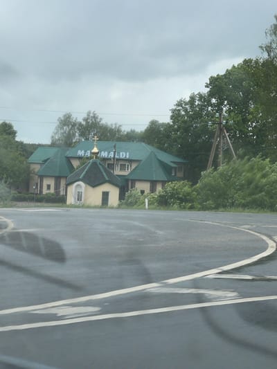 The image shows a view from inside a vehicle looking out onto a road with the "Mama Maldi" building visible. The building has a green roof, a gold cross, and an external sign that says "Mama Maldi" in blue lettering. The location is Кокино, Russia. The sky is overcast. Power lines and trees are also visible. White lines are painted on the road surface. The image is slightly rotated.