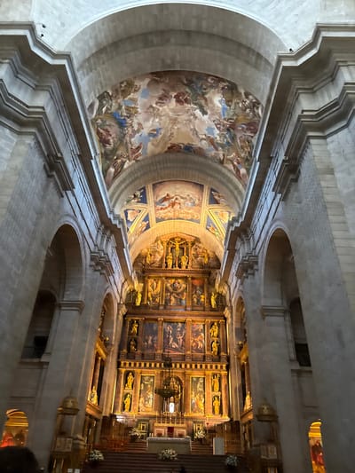 The image depicts the interior of a grand religious edifice, specifically the Basilica within the Royal Monastery of San Lorenzo de El Escorial, located in San Lorenzo de El Escorial, Spain. The perspective is from the nave, looking directly towards the high altar and reredos.

The environment is characterized by massive, light-colored stone architecture, including soaring vaulted ceilings and large, rounded arches. The uppermost vaulted ceiling features an extensive fresco painting depicting numerous celestial figures, angels, and divine beings against a background of clouds and sky. Below this, a secondary, smaller vaulted section, directly above the reredos, is adorned with additional frescoes displaying religious figures and scenes, illuminated by a warm, artificial light source.

The focal point is the monumental, multi-tiered reredos (altarpiece) constructed from dark wood with intricate gilded ornamentation. The reredos is densely populated with numerous framed paintings depicting various religious narratives, interspersed with golden statues of saints or biblical figures housed in niches. At the apex of the reredos, a prominent crucifix with a sculpture of Christ is centered, flanked by more golden statues.

Below the reredos, a white altar table is visible, adorned with white floral arrangements and several candlesticks. A large, ornate chandelier hangs centrally above the altar. Multiple wide steps, covered in a dark material, lead up to the altar area, also featuring additional white floral arrangements. A low, dark barrier or railing separates the altar steps from the main floor of the nave.

The nave's side walls are lined with imposing stone pillars and arches, suggesting access to side aisles or chapels. A dark railing, possibly part of an upper gallery or balcony, is visible through an archway on the left. In the lower left foreground, a dark, indistinct silhouette of a person's head and shoulders is visible, indicating an observer of the scene. No specific actions or interactions are discernible beyond this implied observation.