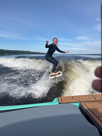 A blonde woman wearing a black wetsuit is wakesurfing on a dark body of water under a bright blue sky with scattered white clouds. She is standing on a white wakesurf board, which is creating a large wake behind a boat, parts of which are visible in the foreground. The woman is looking towards the camera and has her left hand raised in a thumbs-up gesture. The environment is a lake with a tree-lined shore in the distance. This activity is occurring on Lake Gromovskoye, located in the city of Gromovo, Russia.