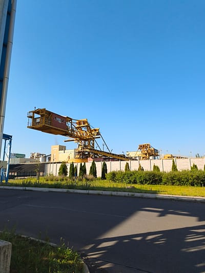 An outdoor, full-shot photograph, taken from ground level, shows a bright, clear, blue sky dominating the upper half of the frame. In the foreground, a dark asphalt road occupies the bottom third, with a concrete curb and some overgrown green grass on the left side. The road is partially covered in the shadows of a large structure.
The middle ground of the image features a row of evenly spaced, conical green trees, behind which is a textured, light-colored concrete wall. Beyond the wall, a complex industrial facility is visible. The most prominent feature of this facility is a large, yellow gantry crane, extending horizontally from left to right across the frame, with its cabin on the far left. Behind the gantry, further industrial buildings with cream-colored walls and darker roofs can be seen, some with visible chimneys emitting a small amount of white smoke. To the far left of the frame, a tall, cylindrical blue structure with metallic accents is partially visible.
The scene appears to be a sunny day in an industrial area. The specific location is the Volevčice mining area, Czechia, which is known for its industrial operations. No people are visible in the image.
