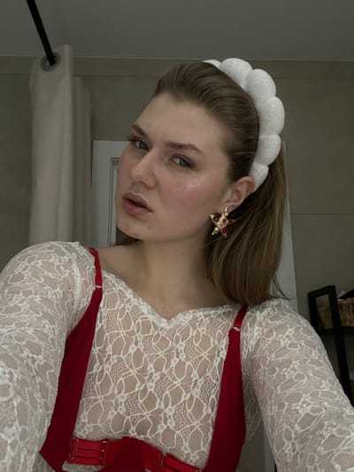 A woman with light skin, light brown hair in a ponytail, and blue eyes looks into the camera. She wears a white, puffy headband and star-shaped earrings. She is wearing a white lace top with a red harness. The background includes a wall, a shelf with objects on it, and a window with a beige curtain. The image is rotated, with the subject appearing at an angle.