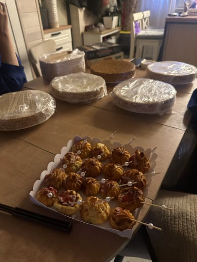 The image depicts a table laden with food items, likely in a domestic setting in Тумілавіцкі сельскі Савет, Belarus. In the foreground, a white scalloped tray contains an assortment of small, golden-brown pastries, some topped with red jam and decorative white pearls. These pastries are skewered with wooden toothpicks, also adorned with pearls. A black utensil rests to the left of the tray. In the background, several round cake bases are stacked and wrapped in clear plastic. One unfrosted cake base is visible without plastic. A person's arm, wearing a dark blue garment, is partially visible on the left side of the image. The table is made of wood, and the environment suggests an indoor scene, possibly a kitchen or dining area, with furniture and cabinets visible in the blurred background. The lighting is dim, creating a warm, ambient atmosphere.