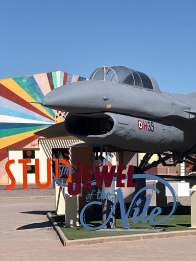 An outdoor scene at Atlas Studios in Ouarzazate, Morocco, features a grey fighter jet replica, elevated on a display stand, under a clear blue sky. The jet exhibits a transparent cockpit canopy, a pointed nose cone, and an intake beneath the cockpit. Its fuselage is marked with an Egyptian Air Force roundel and the alphanumeric designation "OH39." In the background, a building facade is painted with a vibrant, multi-colored radial stripe pattern. Large orange letters spelling "STUDIO" are visible to the left. In the foreground, a sign composed of large red letters reads "The JEWEL" and blue cursive letters spell "of the Nile." The ground consists of light-colored paving stones with a narrow strip of artificial green turf along a curb. Strong direct sunlight casts distinct shadows across the scene. No individuals are present.