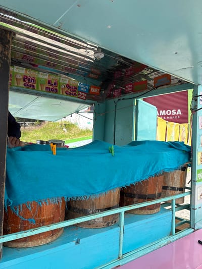 The image depicts the interior of a blue and pink ice cream cart, likely located in Tepeji de Ocampo, Mexico, as suggested by the signs written in Spanish. The cart features a freezer with various flavors of ice cream, including "Pinon", "Limon," "Queso con Coco," "Galleta Oreo," "Mamey," and "Frutos Rojos." Signs for "Mango Maracuya" and "Pico de Gallo" are also visible.

To the right, a blue cloth secured with clothespins partially obscures several wooden barrels that are likely used for storage or as part of the cart's structure. Outside the cart, there is a glimpse of green grass and possibly a building. The view is oriented from a low angle, looking up into the cart. The photograph is taken with the cart in focus.