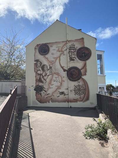 The image captures a building exterior in Weymouth, United Kingdom, featuring a large mural. The mural depicts a historical map of Weymouth Bay, embellished with illustrations and decorative elements. Three coin-like medallions are integrated into the mural's design, with additional drawings depicting a sailing ship, a compass rose, and figures of historical characters. The mural is attributed to "ARTBY ROO.COM," as indicated by text in the lower right corner. Text within the mural identifies landmarks and locations around Weymouth Bay, such as "WEYMOUTH," "smallmouth Sand," and "Portland Ferry." The building is adjacent to a pedestrian walkway, as evidenced by the railings and walkway surface in the foreground. The sky is visible in the background, with a few clouds. A bare tree is visible in the lower left of the image. The lighting suggests that the photograph was taken during the day.