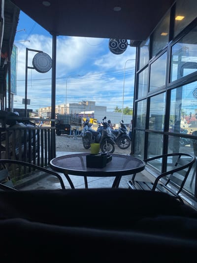 The image captures a view from inside a cafe or restaurant in Keudeublang, Indonesia, looking outwards. In the foreground, a dark metal table and chairs are visible, with a black cup and a yellow cup on the table. Through the large glass windows, a street scene unfolds. Several motorcycles are parked outside, and a small truck is driving on the road. Buildings and power lines are present in the background. Two circular signs are visible outside. One, on the left, says "DOCTOR PENY CLINIC" and the other, on the right, says "68 COFFEE SHOP PREMIUM". The sky is blue with scattered clouds. The overall impression is of a typical street view in a small Indonesian town, seen from the perspective of a shaded establishment.