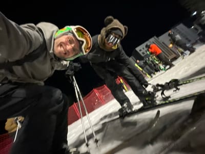 The image depicts a nighttime scene on a snow-covered ski slope in Jahorina, Bosnia and Herzegovina. In the foreground, two individuals are positioned on skis, facing the camera. The subject on the left, male, wears a grey hooded jacket, black pants, a green and white ski helmet with orange goggles pushed up, and eyeglasses, smiling with teeth visible. He holds ski poles in both hands, positioned downwards. The subject on the right wears a dark jacket, dark pants, a brown beanie with bear ears, and horizontally striped ski goggles. This individual's gender is indeterminate. Both subjects are equipped with skis and boots. A red safety net/barrier runs alongside the left edge of the slope behind the foreground subjects. In the mid-ground and background, several other individuals in ski gear are visible, including one person wearing an orange jacket, standing on skis. A light-colored building is present in the distant background to the right. The scene is illuminated by artificial lighting, creating shadows on the snow. The sky is dark.