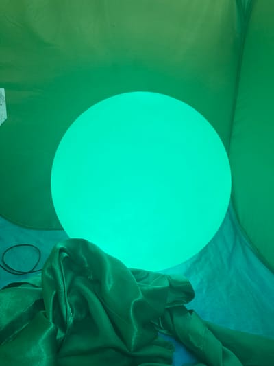 This media file shows a close-up, overhead view of a large, glowing orb emitting a bright, aqua-green light. The orb is positioned in the center-left of the frame and appears to be the primary light source, casting a strong glow that illuminates the surrounding environment. The scene is enclosed, possibly within a tent or a similar fabric structure, as indicated by the draped, translucent green and teal fabric that fills the right and top portions of the image. The fabric has a soft, silky sheen and is gathered in folds, creating a dynamic texture. To the left of the orb, the fabric appears more matte and is a lighter shade of green, suggesting a gradient of color or different lighting conditions within the enclosed space. A black cord is visible trailing from the bottom right corner, disappearing into the fabric. There is a small, white rectangular label or piece of paper visible at the bottom left edge of the frame, partially obscured by the green fabric, with some faint text printed on it. The overall impression is one of soft, diffused light and a somewhat abstract, atmospheric setting. No people or specific landmarks are visible, and the time of day and weather are not discernible from the image. The location context is Wakefield, United Kingdom, but this is not visually evident within the image itself.