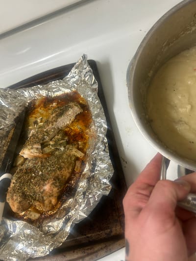 The scene depicts a kitchen environment in the city of Reno, United States of America. A white stovetop surface with visible burner elements occupies the background. On the left, a dark baking sheet holds crumpled aluminum foil containing a cooked piece of seasoned poultry or fish. This food item is heavily coated with green herbs and red flakes, with one end cut or broken, revealing its white interior. Cooked potato or onion pieces are intermingled with a brown liquid on the foil. A black-handled serving utensil with a silver head rests on the aluminum foil to the left of the food. On the right, a silver metal saucepan containing a light-colored, creamy, semi-liquid food with visible dark and faint red specks is positioned on the stovetop. A human hand, appearing to be an adult's, grasps the handle of this saucepan. The hand has a dark, indistinct tattoo on its dorsal wrist/forearm area.