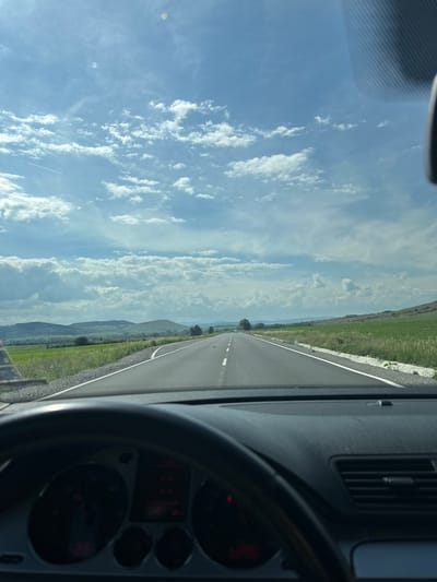 The image captures a driver's perspective from inside a car, looking forward down a road on a bright, sunny day. The scene is primarily focused on the open road and the natural landscape.

In the foreground on the right side, elements of the car's interior are visible, including a dark grey dashboard, an air vent with vertical slats, and a portion of the steering wheel. Behind the steering wheel, the instrument cluster shows some faint red indicator lights, but no specific text or readings are discernible. A patterned grey fabric, possibly a sun visor or the car's headliner, is visible in the top left of the original image (top right after mental rotation).

The main subject is a straight, two-lane asphalt road, marked with a solid white line along its edge, stretching into the distant horizon. On both sides of the road, lush green fields extend outwards, featuring various vegetation and hints of wildflowers, possibly red poppies, near the shoulder. Beyond the fields, a chain of hazy blue-grey hills or mountains creates a natural backdrop.

Above the landscape, a vast blue sky is filled with scattered, fluffy white cumulus clouds, particularly concentrated on the left side. A thin white contrail, likely from an aircraft, is visible in the lower left portion of the sky. The overall setting suggests a daytime drive through a rural or semi-rural area, consistent with the provided location context of Drugan, Bulgaria. The clear sky and bright lighting indicate good weather.
