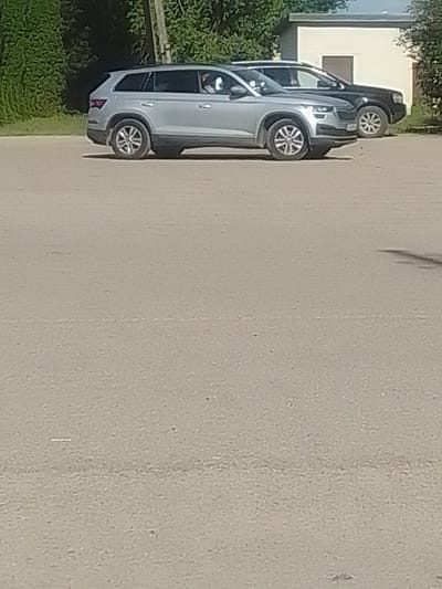 In Koknese, Latvia, two cars are parked side-by-side on an asphalt surface. The car in the foreground is a silver SUV, and it is parked facing right. The car in the background is a black SUV, and it is parked facing right. Behind the cars, there is a building and some trees. The sky is visible above the trees.