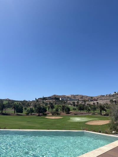 The image depicts a scenic view of a golf course in Urbanización Salobre Golf, Spain, with a swimming pool in the foreground. The clear blue sky dominates the upper half of the frame. Below the sky, a green golf course stretches across the middle ground, dotted with palm trees and other vegetation. Several sand traps are visible on the course. In the background, a rocky hillside is topped with white buildings, suggesting a resort or residential area. The swimming pool in the foreground is filled with turquoise water, its surface rippling and reflecting the sunlight. The edge of the pool and a wooden deck are also visible. There are no people in the image. The overall impression is one of a luxurious and tranquil vacation setting.