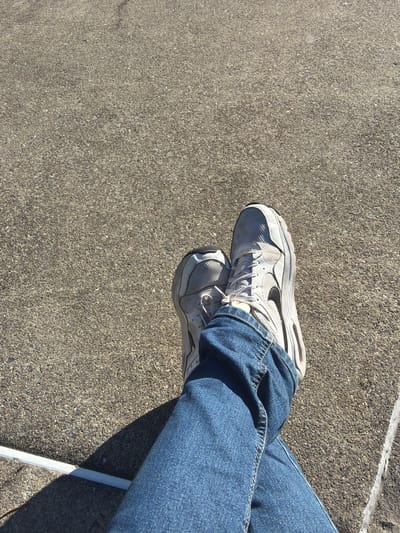 The image shows a person sitting with legs crossed, wearing blue jeans and white athletic shoes with a black swoosh logo, on a concrete surface. The setting appears to be outdoors, with a sidewalk or paved area visible. A white object or edge is seen near the legs. The location is Edison, United States of America.