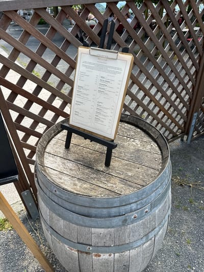This media file features an outdoor seating area, likely at a restaurant or cafe, with a prominent wooden barrel and a menu displayed on an easel. A lattice wooden fence forms a backdrop, with blurry figures of people visible through the gaps, suggesting a public space. The menu, written in German, lists various food and drink options under categories such as "APERITIF," "LES PLATS" (dishes), "ENTRÉE," "POTAGE" (soup), and "DESSERT." The overall setting appears to be relaxed and inviting, possibly during daytime given the natural lighting and shadows. The presence of a barrel suggests a possible winery or a place with a rustic theme. The text on the menu includes items like "Tagliatelle," "Steak Frites," "Wiener Schnitzel," and "Crème Brûlée," along with prices. The restaurant's name, "CHARLES Bistro & Cafe," is clearly visible at the top of the menu, and a welcoming message in German is also present. The overall impression is one of a pleasant outdoor dining experience in a European setting.
