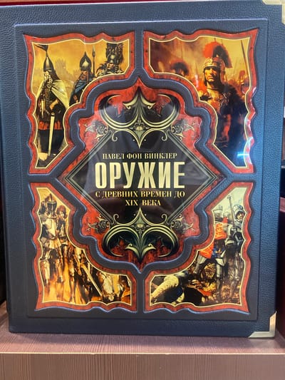 The image shows the cover of a book titled "ОРУЖИЕ С ДРЕВНИХ ВРЕМЕН ДО XIX ВЕКА" (Weapons from Ancient Times to the 19th Century) by ПАВЕЛ ФОН ВИНКЛЕР (Pavel von Winkler). The cover is dark blue and has an ornate design featuring images of armored warriors from different historical periods, framed within red and gold decorative borders. The book is displayed on a wooden shelf. There is no information in the image or its crops to suggest a location in Mikhalkovo, Russia.
