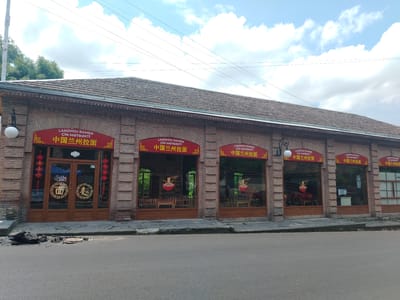 The image depicts the exterior of a brick building in the city of Shaki, Azerbaijan, which houses a restaurant named "LANZHOU RAMEN" (Chinese: 中国兰州拉面). The building has a tiled roof and multiple arched windows. Red banners with the restaurant's name in both English and Chinese are displayed above several of the windows. The entrance features double wooden doors with circular emblems. In front of the building, the asphalt road shows signs of disrepair, with a hole and debris on the pavement. The scene is set on a cloudy day, with power lines visible against the sky. Trees and greenery are visible behind the building.